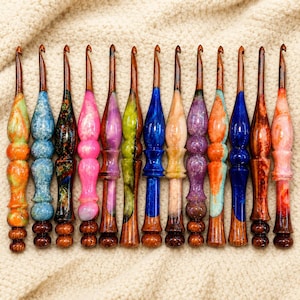 Peut inclure: Une collection de treize crochets disposés en rangée. Chaque crochet présente un manche en résine coloré unique avec un motif marbré ou tourbillonnant, associé à une tige en bois lisse et effilée avec une pointe métallique à l'extrémité.