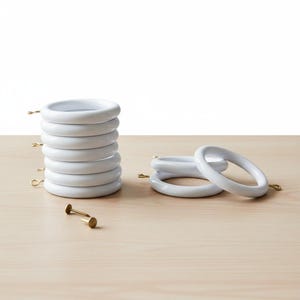 Pode incluir: Um conjunto de argolas brancas para cortinas com ganchos dourados e um prego dourado. Vários anéis estão empilhados, outros estão colocados perto. Os anéis estão sobre uma superfície de madeira clara.