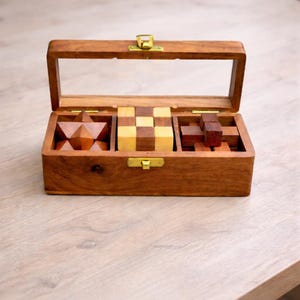 May include: A wooden puzzle box with a clear lid, containing three different wooden puzzles. The box is rectangular with brass-coloured clasps. The puzzles include a star-shaped puzzle, a checkerboard-patterned cube puzzle, and a block puzzle.