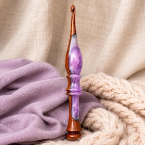 Può includere: Un uncinetto artigianale con manico in legno marrone scuro lucido, integrato con una sezione centrale in resina liscia marmorizzata viola e bianca. L'uncinetto presenta un collo affusolato e una base arrotondata con venature del legno.
