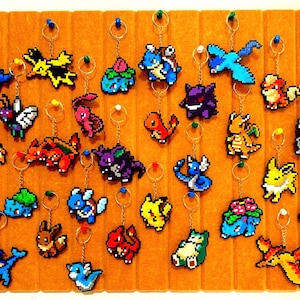 Può includere: Una collezione di portachiavi pixelati di personaggi, ciascuno con un anello e una catena dorati, esposti su una tavola di feltro marrone. I portachiavi presentano varie creature colorate, tra cui draghi e uccelli.