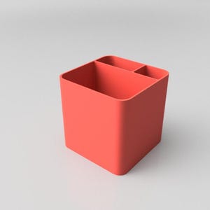 May include: A coral desk tidy with three compartments. The cube-shaped organiser has rounded corners and a smooth, matte finish. The compartments are of varying sizes, suitable for pens, pencils, and other stationery items.