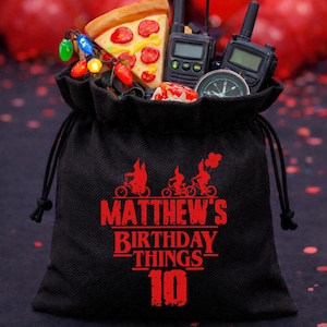 May include: Black drawstring bag with red text that reads "Matthew's Birthday Things 10". The bag is filled with a pizza slice, walkie-talkies, a compass, and string lights. Red balloons are in the background.