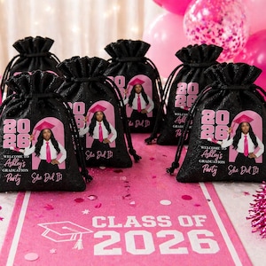May include: Black drawstring favor bags with a pink and white design for a graduation party. Each bag features a photo of a graduate, the year 2026, and the text "She Did It!" The scene includes pink balloons and decorations.