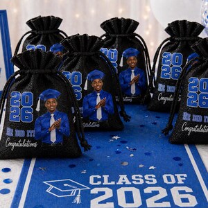 May include: Black drawstring bags with a graduation theme. Each bag features a photo of a graduate, the year "2026", and the text "He DID IT! Congratulations!". The bags are arranged on a table with a blue runner that reads "Class of 2026".