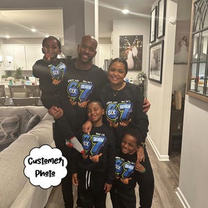 May include: A family of six wearing matching black sweatshirts and sweatpants. The sweatshirts have a blue and green graphic that says "Dillon was SIX, Now he's SEVEN". The family is smiling and posing for a photo.