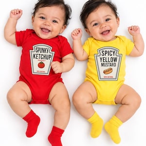 May include: Two short-sleeved baby bodysuits. One is red with a graphic of a tomato and the text SPUNKY KETCHUP, 100% NATURAL. The other is yellow with a graphic of a hot dog and the text SPICY YELLOW MUSTARD, 100% NATURAL. Both include matching socks.
