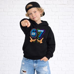 May include: Black hooded sweatshirt with the numbers "67" in blue and green, and the word "SIX" in a circle. The sweatshirt has a front pocket and is worn by a person wearing a black baseball cap and blue jeans.