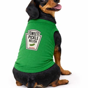 May include: A green dog shirt featuring a graphic of a condiment label that reads SWEET PICKLE RELISH with an illustration of a pickle and the text 100% NATURAL. The shirt is worn by a black and tan dachshund with its head turned toward the camera.