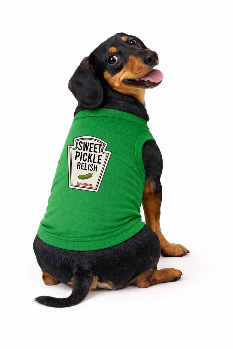 May include: A green dog shirt featuring a graphic of a condiment label that reads SWEET PICKLE RELISH with an illustration of a pickle and the text 100% NATURAL. The shirt is worn by a black and tan dachshund with its head turned toward the camera.