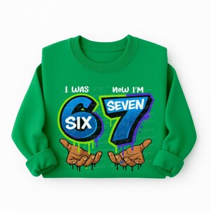 May include: A green sweatshirt with the text "I WAS SIX NOW I'M SEVEN". The number "6" is blue with a green outline, and the number "7" is blue with a purple outline. The sweatshirt also features two hands with brown skin.