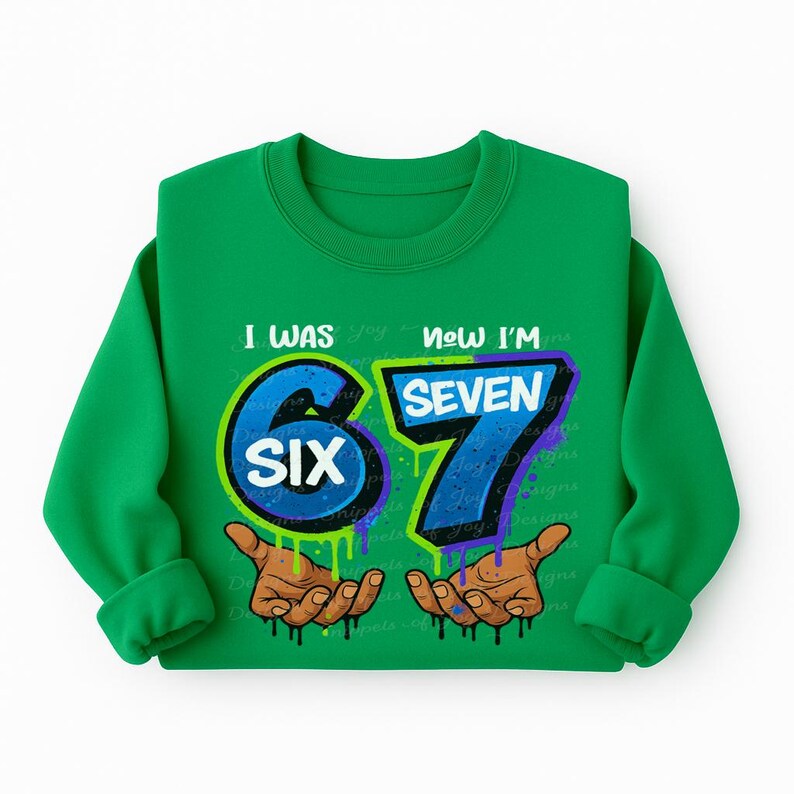 May include: A green sweatshirt with the text "I WAS SIX NOW I'M SEVEN". The number "6" is blue with a green outline, and the number "7" is blue with a purple outline. The sweatshirt also features two hands with brown skin.