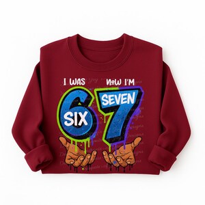 May include: A maroon sweatshirt with the text "I WAS SIX NOW I'M SEVEN" in a graffiti-style design. The numbers 6 and 7 are in blue with green and purple outlines, and the word "SIX" is in white. The design includes brown hands with dripping paint.