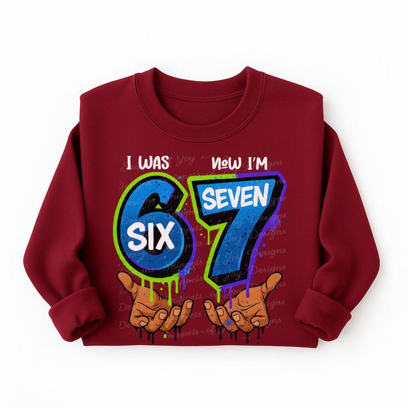 May include: A maroon sweatshirt with the text "I WAS SIX NOW I'M SEVEN" in a graffiti-style design. The numbers 6 and 7 are in blue with green and purple outlines, and the word "SIX" is in white. The design includes brown hands with dripping paint.
