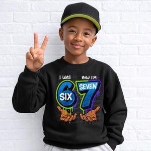 May include: A black sweatshirt with the text "I WAS SIX, NOW I'M SEVEN" in blue and green. The number 6 is in a circle, and the number 7 is in a rectangle. The sweatshirt is worn by a child wearing a black baseball cap.