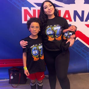 May include: A family of four poses for a photo. The mother and two children are wearing black t-shirts with the number "67" and the word "SIX" and "SEVEN" printed on them. The youngest child is being held by the mother. The background is a blue and white wall.
