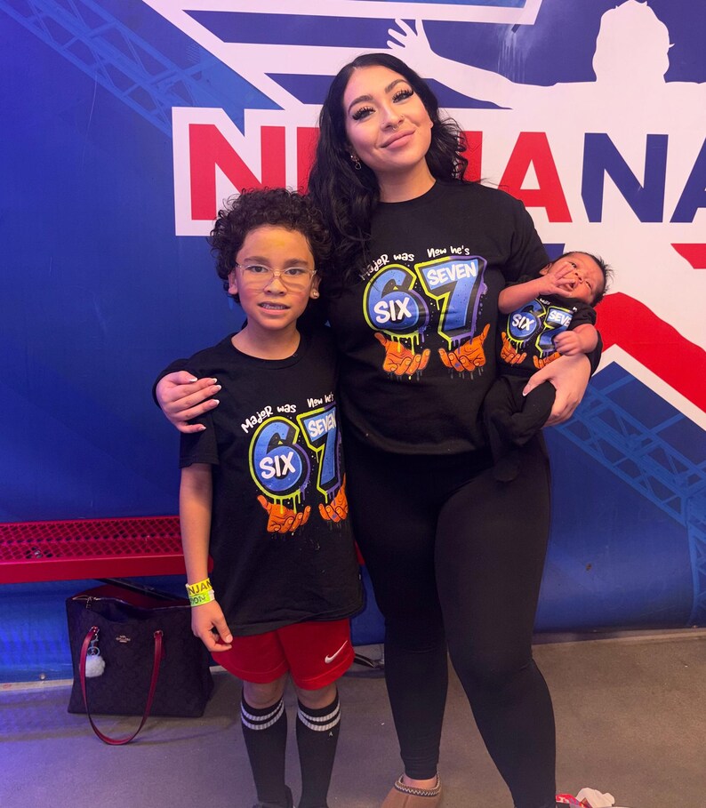 May include: A family of four poses for a photo. The mother and two children are wearing black t-shirts with the number "67" and the word "SIX" and "SEVEN" printed on them. The youngest child is being held by the mother. The background is a blue and white wall.