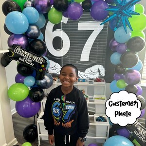 May include: A birthday party scene with a balloon arch in shades of green, purple, blue, and black. The number 67 is displayed prominently. A child is wearing a black sweatshirt with the text "I was SIX, Now I'M EVEN 67".