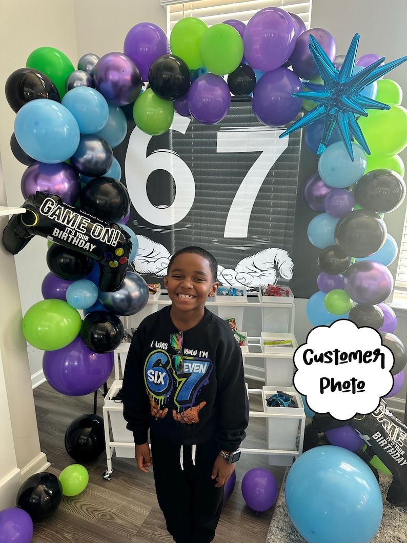 May include: A birthday party scene with a balloon arch in shades of green, purple, blue, and black. The number 67 is displayed prominently. A child is wearing a black sweatshirt with the text "I was SIX, Now I'M EVEN 67".