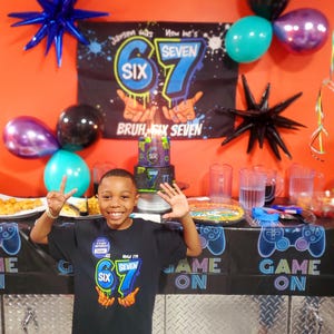 May include: A young boy smiles at his birthday party, wearing a black t-shirt with the text "Now I'm Seven". The party decorations include balloons, a banner, and a cake. The banner reads "Jarsen was Six, now he's Seven".