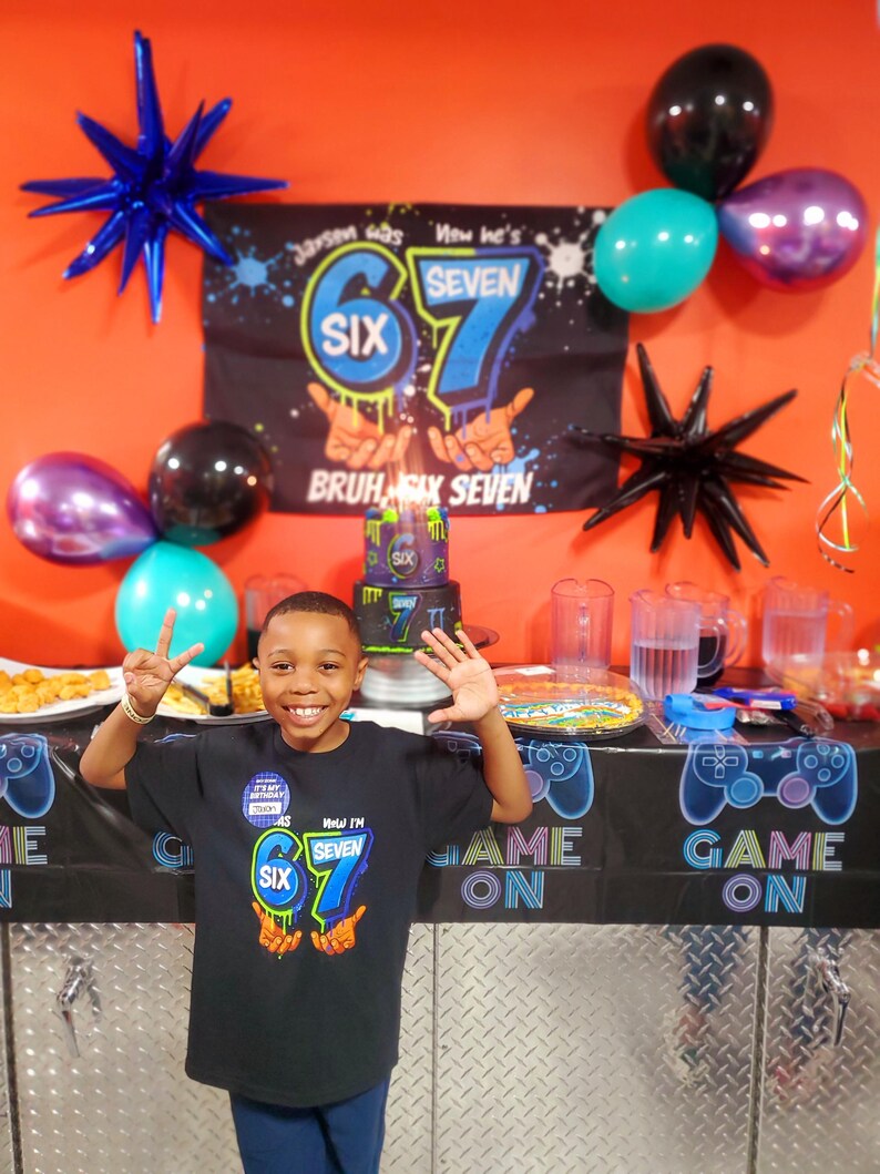 May include: A young boy smiles at his birthday party, wearing a black t-shirt with the text "Now I'm Seven". The party decorations include balloons, a banner, and a cake. The banner reads "Jarsen was Six, now he's Seven".