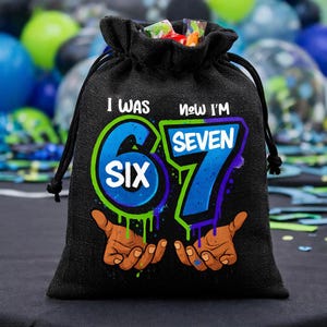 May include: A black drawstring gift bag featuring a graphic print with the text I WAS SIX NOW I'M SEVEN. The design includes large blue numbers 6 and 7 with green and purple outlines, and two open hands reaching upward from the bottom of the bag.