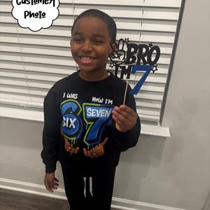 May include: A young person wearing a black sweatshirt and black pants. The sweatshirt has the text "I WAS SIX, NOW I'M SEVEN" with colorful graphics. They are holding a blue and black "BRO I'M 7" birthday sign.