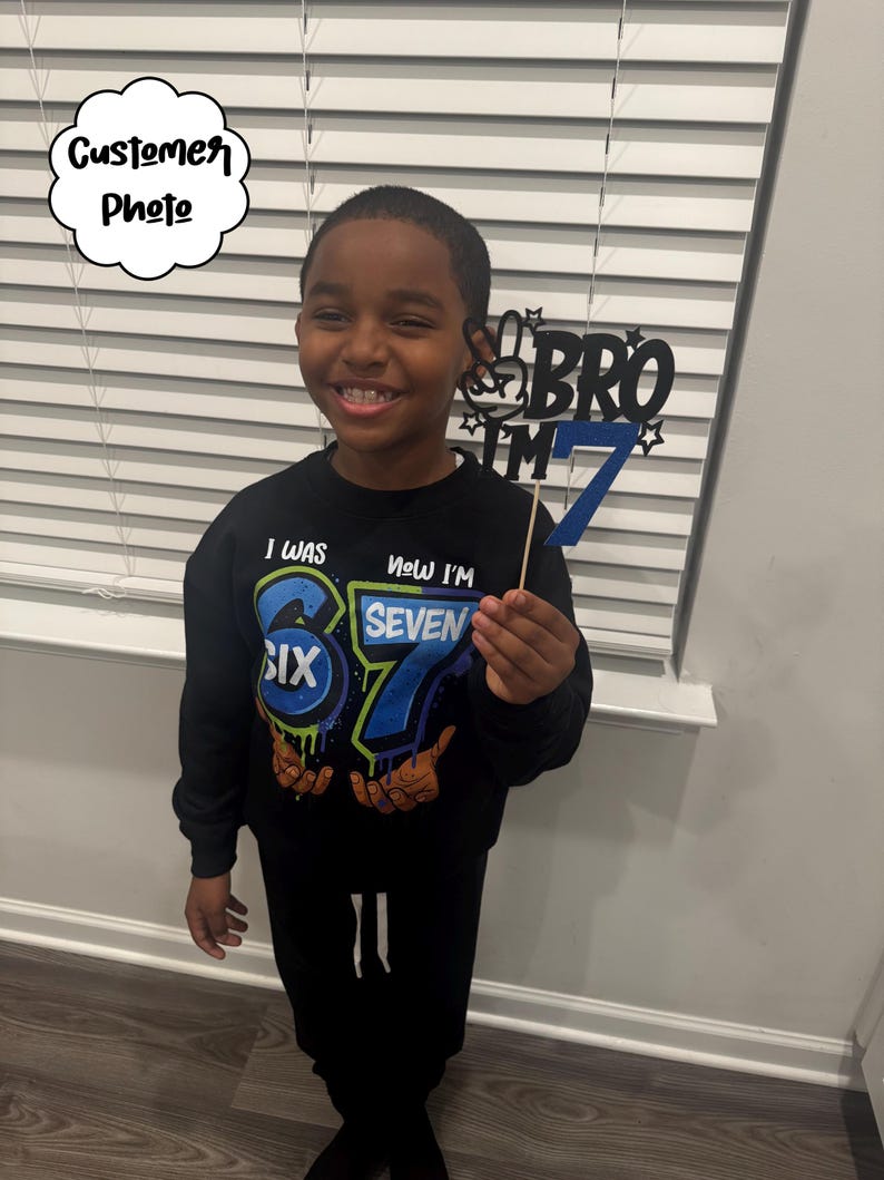 May include: A young person wearing a black sweatshirt and black pants. The sweatshirt has the text "I WAS SIX, NOW I'M SEVEN" with colorful graphics. They are holding a blue and black "BRO I'M 7" birthday sign.