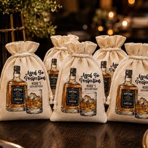May include: A set of beige drawstring fabric favor bags featuring a printed graphic of a whiskey bottle and a glass with ice. The text reads Aged To Perfection and Michael's 50th Birthday. The bags are made of a textured, woven material.