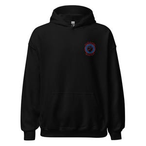 Puede incluir: Sudadera con capucha negra con bolsillo delantero y capucha con cordón. Presenta un pequeño diseño bordado en la parte superior izquierda del pecho. El diseño es un emblema circular con detalles en rojo y azul y texto.