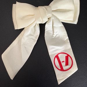 May include: Ivory-colored bow with long, flowing tails. The bow features a large, knotted center and a red circular logo with a stylized "V" and "-" design printed on one tail. The bow is made of a soft, textured fabric.