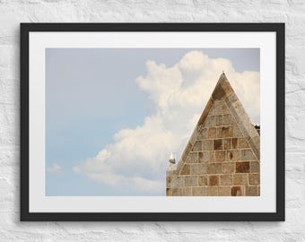 Puede incluir: Fotografía enmarcada de un edificio de piedra con tejado triangular, sobre un cielo azul con nubes blancas y esponjosas. Una gaviota se posa en el borde del edificio. La fotografía está en un marco negro.