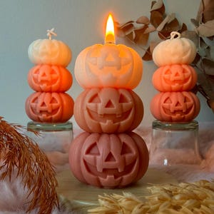Op de afbeelding: Drie stapels van drie uitgesneden pompoenkaarsen in verschillende tinten oranje. De middelste stapel brandt, met een heldere vlam bovenop de bovenste pompoen. Elke pompoen heeft een klassiek jack-o'-lantern gezicht met driehoekige ogen.
