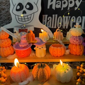 May include: A collection of Halloween-themed candles, including pumpkins, ghosts, and jack-o'-lanterns, arranged on a wooden shelf. Some candles are lit, casting a warm glow. A decorative skull and a sign that says "Happy Halloween" are in the background.