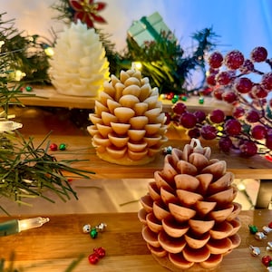 May include: Three pine cone-shaped candles in varying shades of white and brown sit on a wooden shelf, decorated with Christmas greenery and red berries. The candles have a detailed, textured surface, and are lit by a string of lights.