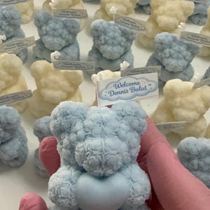 May include: A collection of teddy bear-shaped candles in light blue and cream colors. Each candle has a heart-shaped detail and a small tag that reads "Welcome Dennis Bulut". The candles are arranged on a white surface.