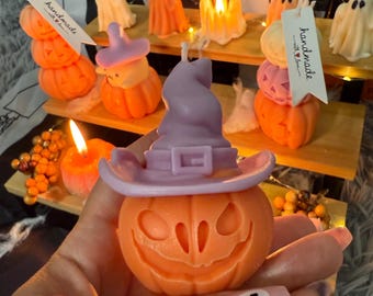 Op de afbeelding: Oranje pompoenvormige kaars met een uitgesneden gezicht, met een paarse heksenhoed. Andere Halloween-kaarsen zijn geesten en pompoenen. De kaarsen worden gepresenteerd op houten planken, met een "handmade with love" label.