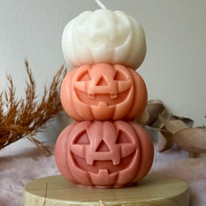 Op de afbeelding: Een gestapelde stompkaars bestaande uit drie uitgesneden pompoenvormen met jack-o'-lantern gezichten. De bovenste pompoen is wit, de middelste is lichtoranje en de onderste is een donkerder terracotta oranje. Een enkele witte lont steekt uit de bovenkant.