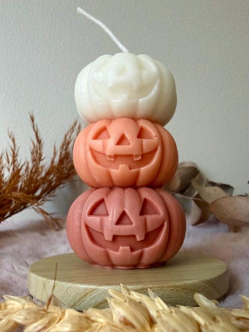 Op de afbeelding: Een gestapelde stompkaars bestaande uit drie uitgesneden pompoenvormen met jack-o'-lantern gezichten. De bovenste pompoen is wit, de middelste is lichtoranje en de onderste is een donkerder terracotta oranje. Een enkele witte lont steekt uit de bovenkant.