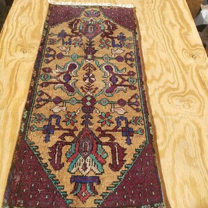 May include: A rectangular, hand-woven rug with a central tan field and intricate geometric patterns in shades of burgundy, teal, and gold. The rug has a burgundy border and white fringe.