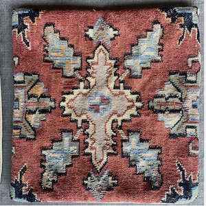 May include: Square, patterned cushion with a red background and a central geometric design in beige, blue, and black. The cushion has a woven texture, suggesting a rug-like appearance, and is likely used for seating or decoration.