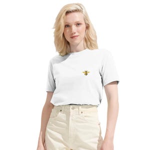 Può includere: T-shirt bianco con un piccolo disegno di ape ricamato sul petto. L'ape è gialla e nera. La maglietta è a maniche corte e ha uno scollo tondo. La modella indossa pantaloni chiari.