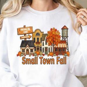 May include: A white long-sleeve sweatshirt featuring a graphic of a small town street with autumn trees, pumpkins, and a water tower. The graphic includes a wooden sign that reads Custom Zip code and the text Small Town Fall in orange letters.