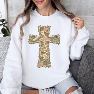 May include: White sweatshirt featuring a large cross design. The cross is decorated with a camouflage pattern in shades of green, brown, and tan. The sweatshirt has a crew neck and long sleeves.