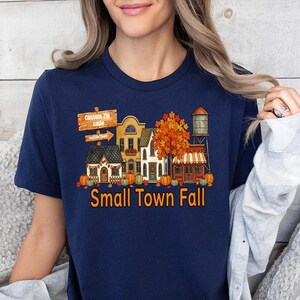 May include: A navy blue t-shirt featuring a graphic of a small town street scene during autumn. The design includes buildings, a water tower, orange trees, and pumpkins. The text reads "Small Town Fall" in orange letters below the illustration.