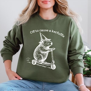 May include: Olive green sweatshirt with the text "Off to cause a kerfuffle". The graphic shows a white illustration of a rodent wearing a witch hat and riding a scooter. The sweatshirt is a casual style.