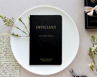 May include: A black officiant book with gold lettering on a white plate. The book says "Officiant" and "Rev. Ethan Miller". The book also has the date "August 21, 2023" and the names "Tristian & Celeste".