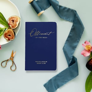 Puede incluir: Un libro de oficiante azul marino con letras doradas. El libro dice "Officiant - Rev. Ethan Miller - Tristan & Delilah August 28, 2021".