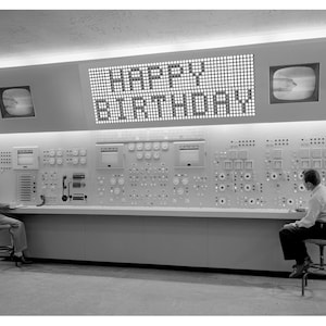 May include: A black and white photo of a control room with two people sitting at a large control panel. The panel has many buttons, switches, and dials. A large screen above the panel displays the text "HAPPY BIRTHDAY".