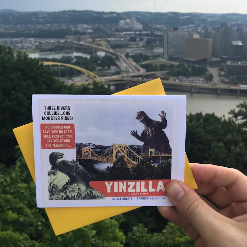 May include: A vintage movie poster for a fictional film called "Yinzilla" with the tagline "Three Rivers Collide...One Monster Rises!" The poster features a giant monster attacking a bridge in a city skyline. The poster is in a hand holding it up against a real city skyline.